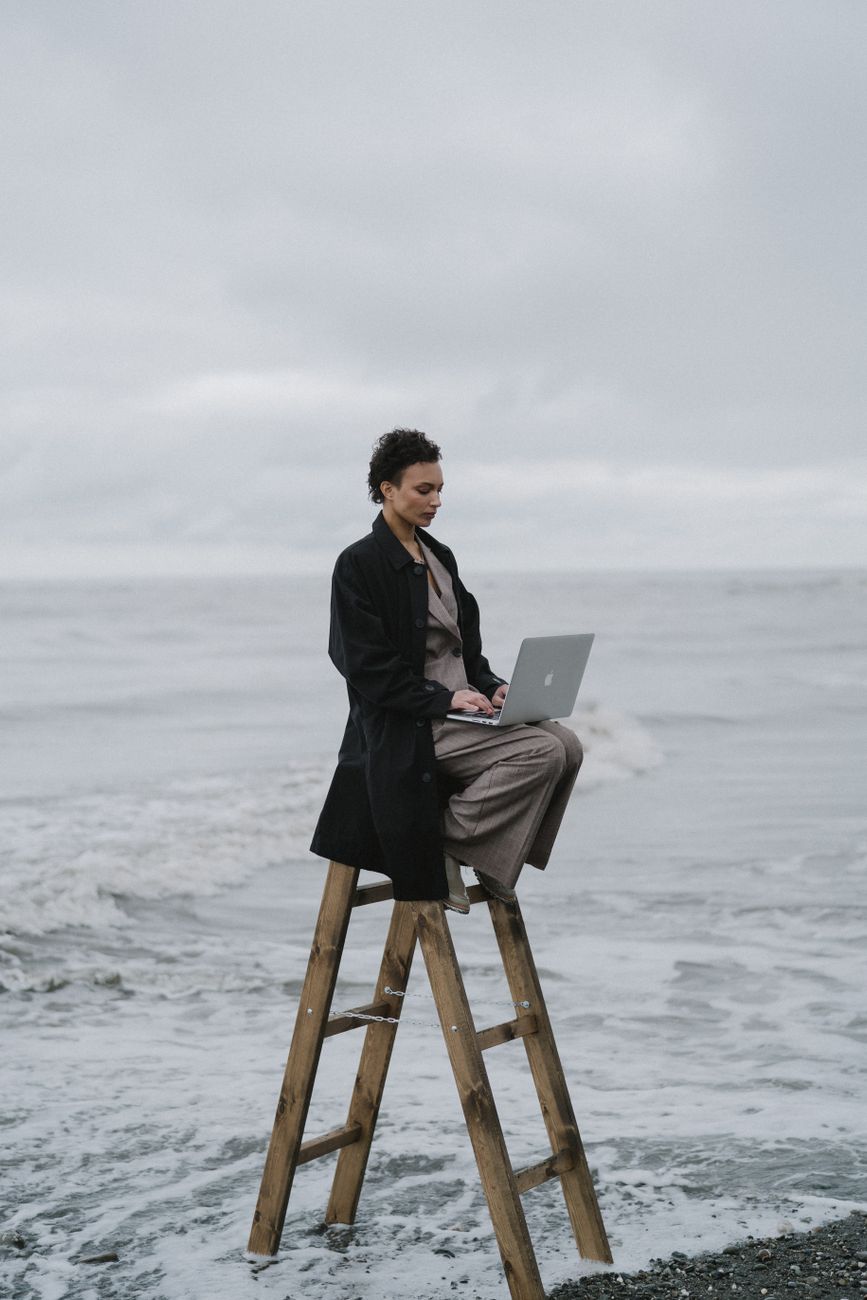 a woman sitting on a wooden ladder at the beach while using her laptop